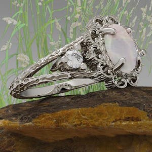 May include: A silver ring with an oval, light-colored gemstone and intricate filigree detailing. The ring is set on a textured rock, with soft green and white plants in the background. The ring's design includes smaller accent stones.