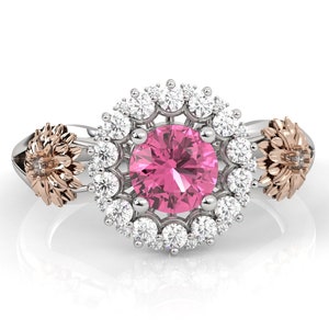May include: A silver ring with a large pink gemstone center, surrounded by a halo of white gemstones. The ring features two gold-colored sunflower accents on either side of the center stone. The ring is set against a white background.