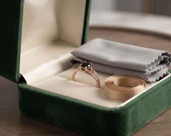 May include: A rose gold ring with a dark round gemstone sits inside a dark green velvet jewelry box. The box contains a folded light gray microfiber cleaning cloth and a small coil of beige ribbon next to the ring on a cream lining.