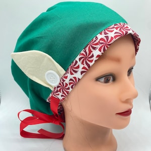 May include: A green scrub cap with a white button and a cream-colored elf ear attached. The cap has a red and white peppermint candy print trim and a red ribbon tie. The cap is on a mannequin head.