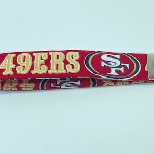 Puede incluir: Llavero rojo y dorado con las palabras "49ERS" y un logotipo rojo y dorado con las letras "SF".