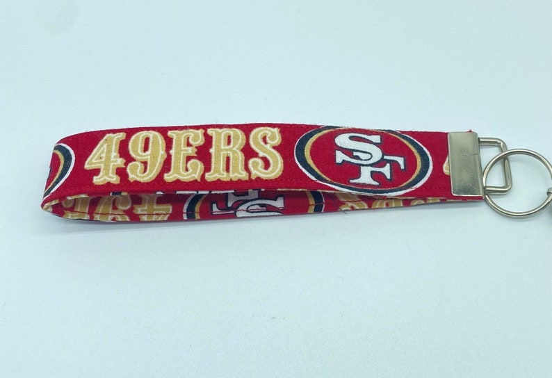 Puede incluir: Llavero rojo y dorado con las palabras "49ERS" y un logotipo rojo y dorado con las letras "SF".