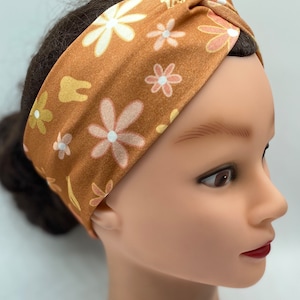 May include: A brown turban headband with a floral pattern featuring white and pink flowers. The headband is made of a soft, stretchy fabric and has a twisted knot design.