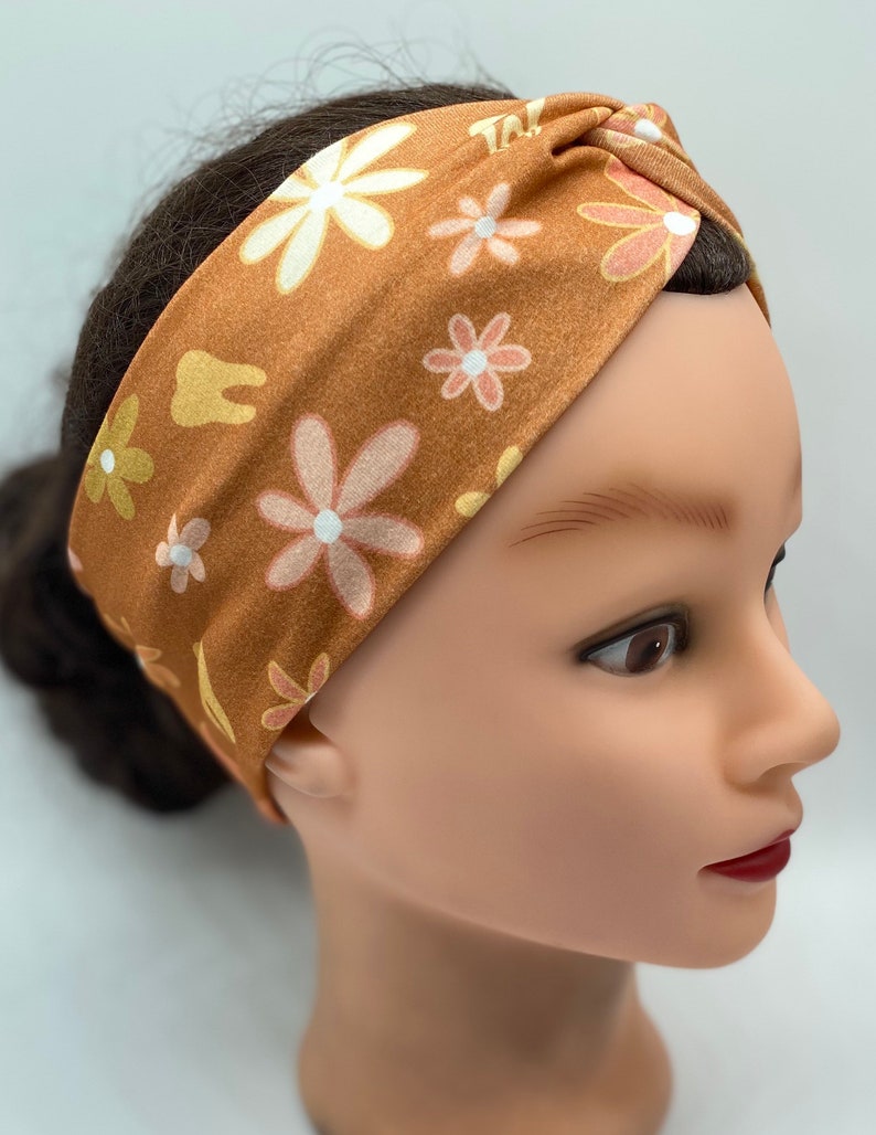 May include: A brown turban headband with a floral pattern featuring white and pink flowers. The headband is made of a soft, stretchy fabric and has a twisted knot design.