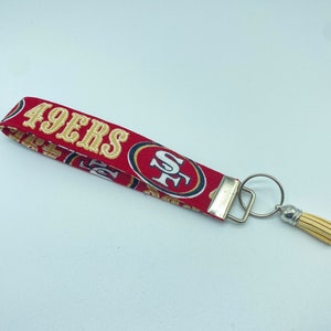 Puede incluir: Llavero de tela rojo y dorado con borla y anilla. La tela presenta el logotipo de los San Francisco 49ers y las palabras "49ers".