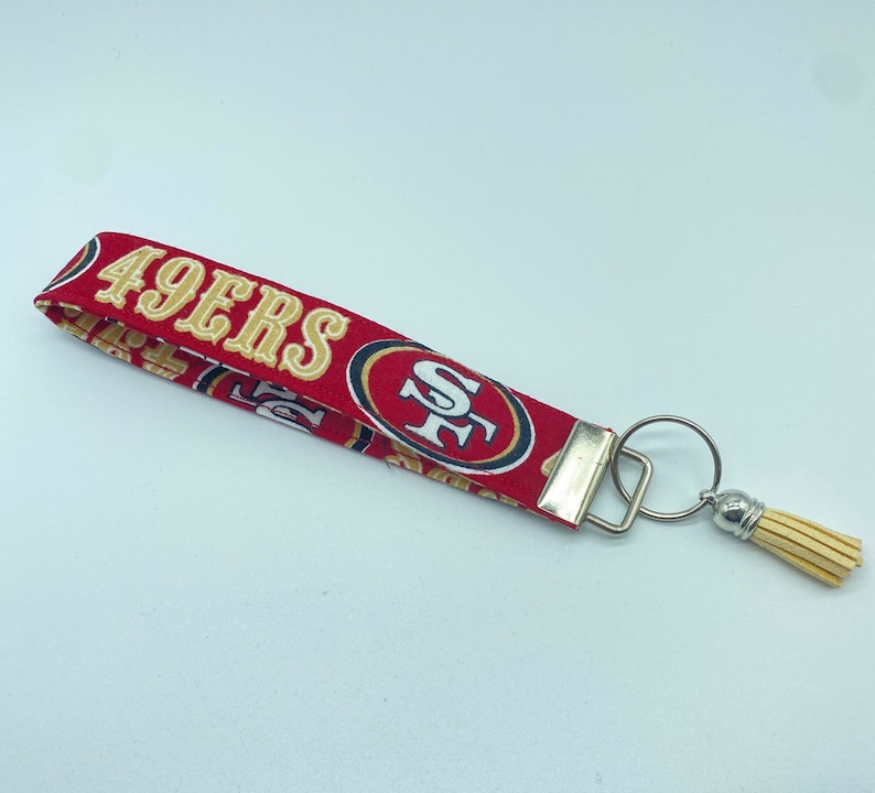 Puede incluir: Llavero de tela rojo y dorado con borla y anilla. La tela presenta el logotipo de los San Francisco 49ers y las palabras "49ers".