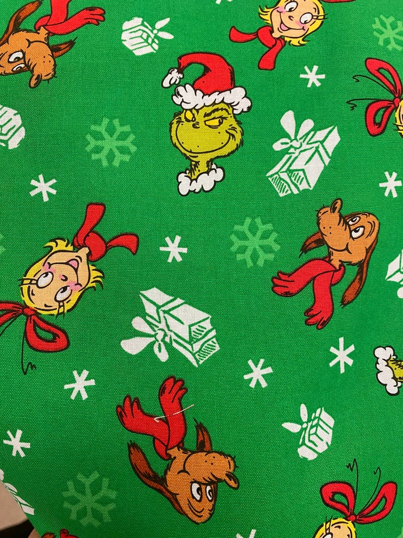 Może przedstawiać: Zielony materiał z powtarzającym się wzorem Grincha, psa Maxa, Cindy Lou Who, płatk&oacute;w śniegu i prezent&oacute;w. Grinch ma na sobie czapkę Świętego Mikołaja i ma złośliwy uśmiech. Pies Max ma na sobie czerwony szalik. Cindy Lou Who ma na sobie czerwoną kokardkę. Prezenty są białe z czerwoną kokardką.