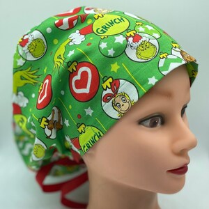 Może przedstawiać: Zielony czepek chirurgiczny z nadrukiem świątecznym przedstawiającym Grincha, Cindy Lou Who i ozdoby. Czepek ma czerwone sznurki.
