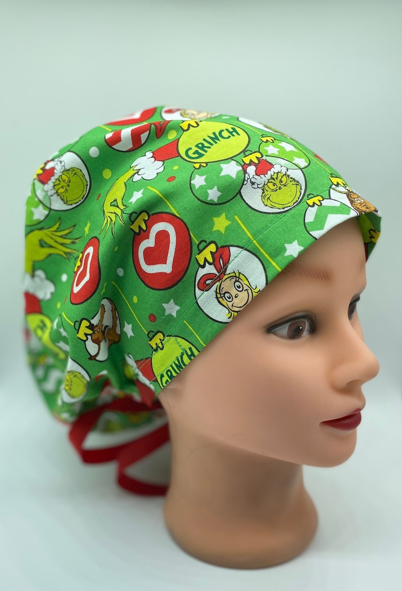 Może przedstawiać: Zielony czepek chirurgiczny z nadrukiem świątecznym przedstawiającym Grincha, Cindy Lou Who i ozdoby. Czepek ma czerwone sznurki.