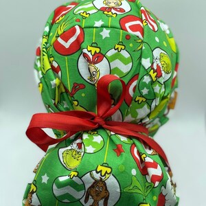 Może przedstawiać: Zielony chirurgiczny czepek z tkaniny z nadrukiem świątecznym przedstawiającym Grincha, Cindy Lou Who i ozdoby. Czepek jest związany czerwoną wstążką.