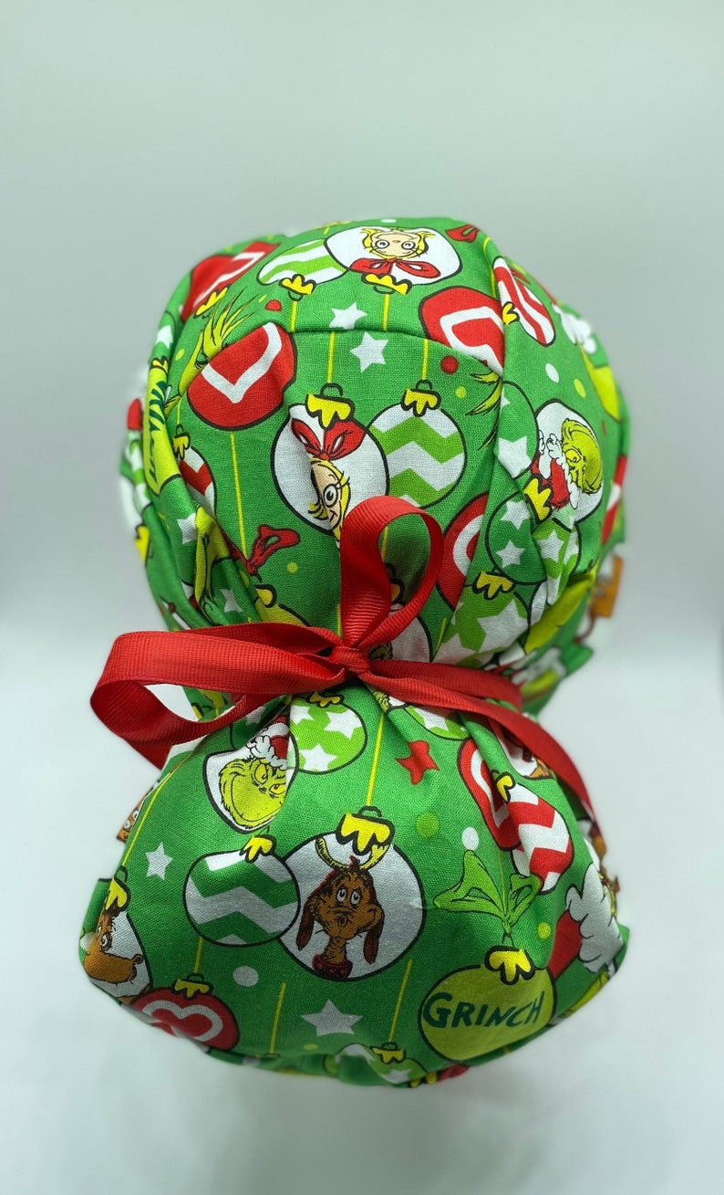 Może przedstawiać: Zielony chirurgiczny czepek z tkaniny z nadrukiem świątecznym przedstawiającym Grincha, Cindy Lou Who i ozdoby. Czepek jest związany czerwoną wstążką.