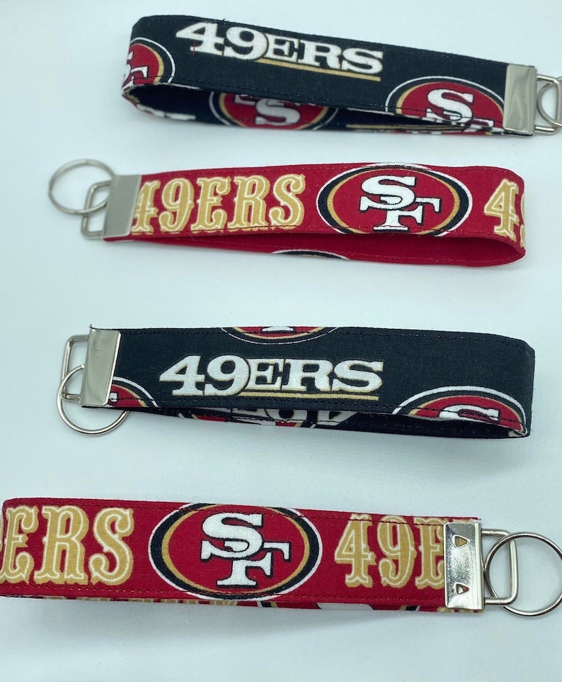 Puede incluir: Cuatro llaveros diferentes con el logotipo de los San Francisco 49ers. Los llaveros son rojos, negros y dorados con el logotipo del equipo y las palabras "49ers" impresas en ellos.