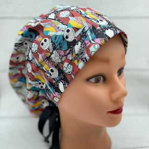 May include: A patterned scrub cap featuring characters from The Nightmare Before Christmas. The cap has a black ribbon tie and is displayed on a mannequin head. The fabric design includes Jack Skellington, Sally, and other characters in a colourful pattern.