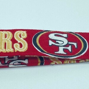 Puede incluir: Llavero de tela rojo, blanco y dorado con borla. La tela tiene las palabras "49ERS" y un logotipo con las letras "SF".