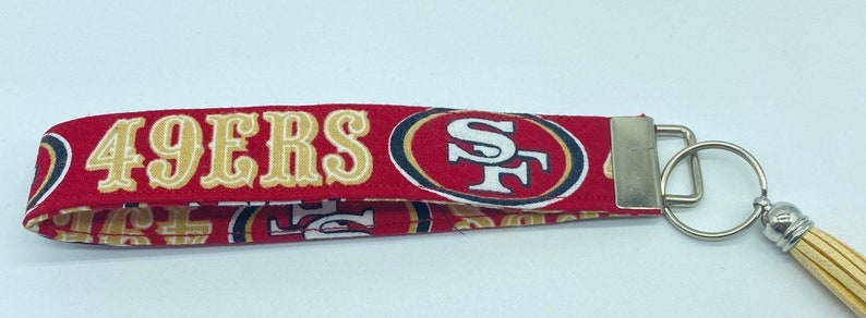 Puede incluir: Llavero de tela rojo, blanco y dorado con borla. La tela tiene las palabras "49ERS" y un logotipo con las letras "SF".