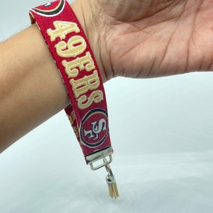 Puede incluir: Pulsera de tela roja y dorada con el texto "49ERS" y un logotipo dorado de los San Francisco 49ers. La pulsera tiene un llavero plateado con un borla.