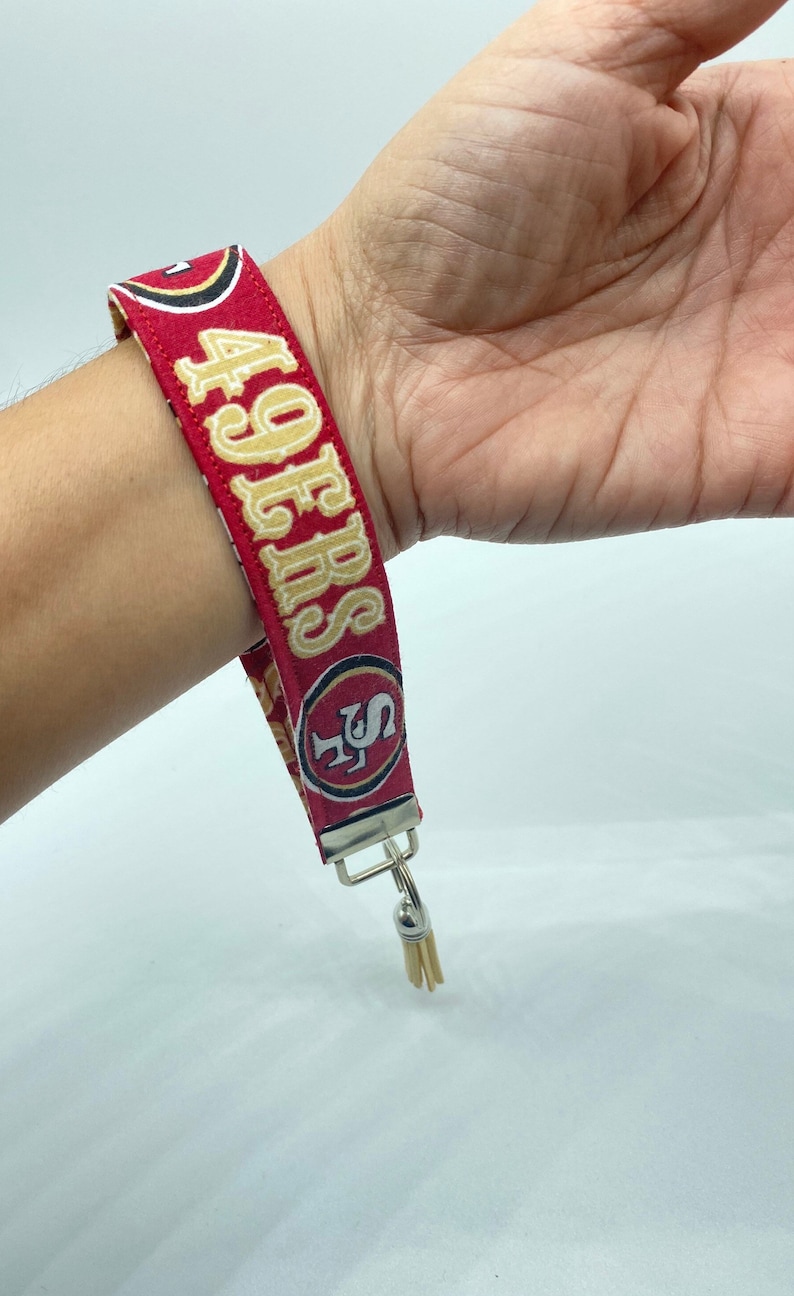 Puede incluir: Pulsera de tela roja y dorada con el texto "49ERS" y un logotipo dorado de los San Francisco 49ers. La pulsera tiene un llavero plateado con un borla.