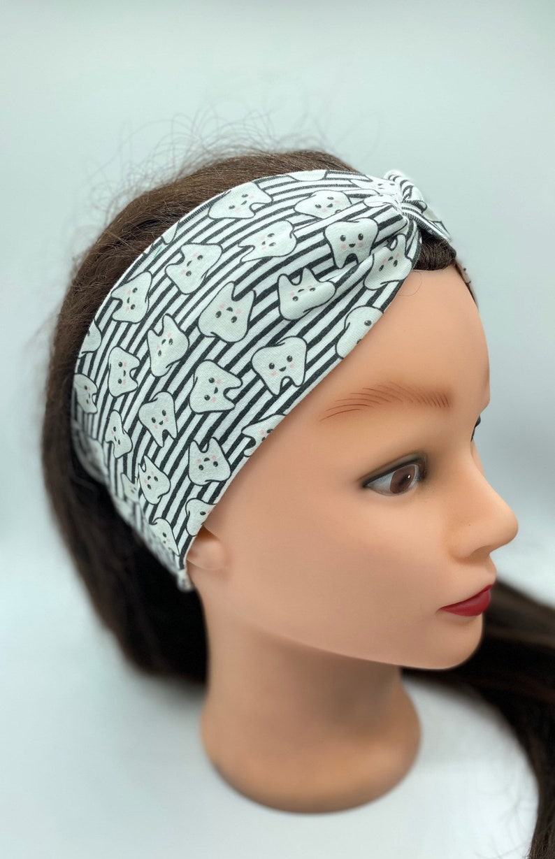 May include: A white and black patterned headband with a design of cartoon teeth. The headband is made of a soft, stretchy fabric and is designed to be worn as a turban.