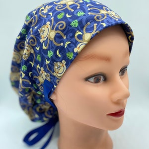 May include: A blue scrub cap with a repeating pattern of cartoon monkeys, bananas, and green leaves. The cap has a blue band and ties. The cap is on a mannequin head.