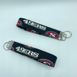 Puede incluir: Dos llaveros negros con un logotipo de los 49ers en rojo y dorado y el texto "49ers" impreso en ellos. Cada llavero tiene un anillo de llave plateado.