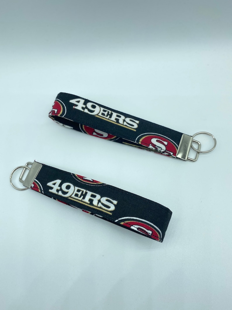 Puede incluir: Dos llaveros negros con un logotipo de los 49ers en rojo y dorado y el texto "49ers" impreso en ellos. Cada llavero tiene un anillo de llave plateado.