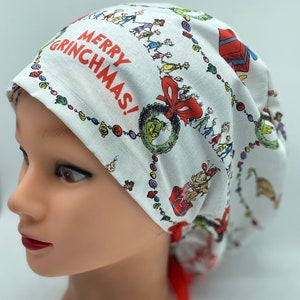 May include: White scrub cap featuring a festive Grinch-themed print. The cap displays the text "MERRY GRINCHMAS!" in red, surrounded by Grinch characters, gifts, and ornaments. A red ribbon is tied at the back.