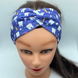 May include: A blue headband with a white tooth pattern. The headband is twisted and has a white tooth with a pink bow on it.