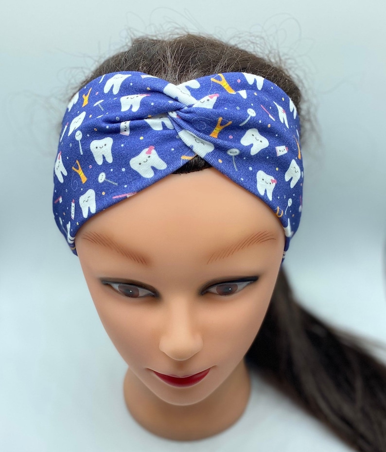 May include: A blue headband with a white tooth pattern. The headband is twisted and has a white tooth with a pink bow on it.