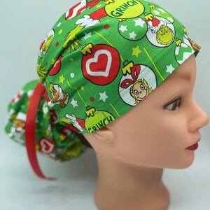 Może przedstawiać: Zielony chirurgiczny czepek scrub z nadrukiem świątecznym przedstawiającym Grincha, Cindy Lou Who i ozdoby. Czepek ma czerwoną wstążkę, a widoczny jest napis "Grinch".