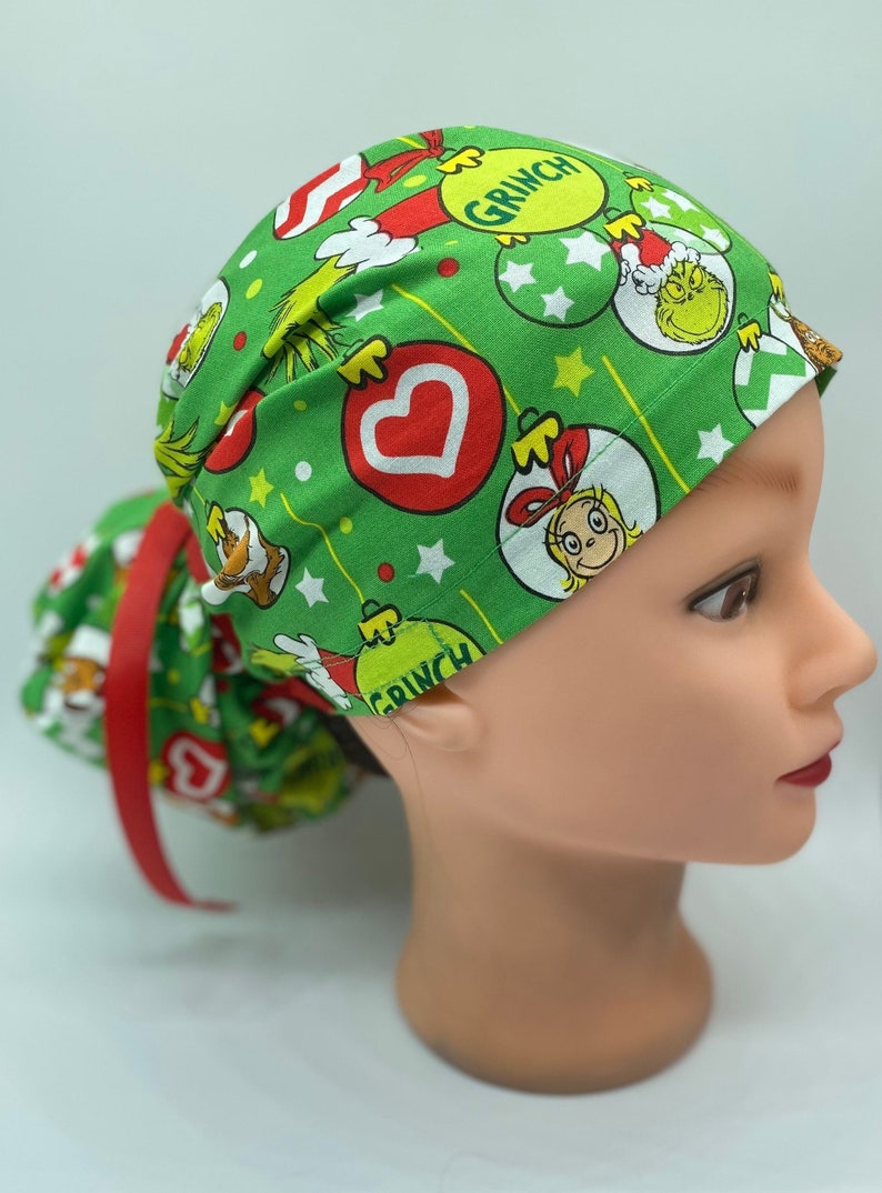Może przedstawiać: Zielony chirurgiczny czepek scrub z nadrukiem świątecznym przedstawiającym Grincha, Cindy Lou Who i ozdoby. Czepek ma czerwoną wstążkę, a widoczny jest napis "Grinch".