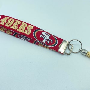 Puede incluir: Llavero de tela rojo y dorado con borla. La tela presenta el logotipo de los San Francisco 49ers y las palabras "49ers".