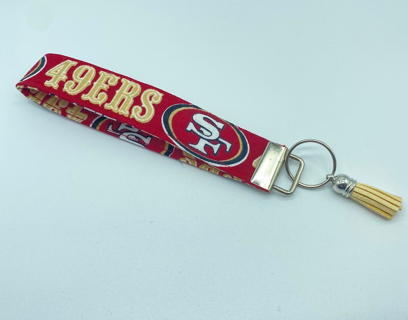 Puede incluir: Llavero de tela rojo y dorado con borla. La tela presenta el logotipo de los San Francisco 49ers y las palabras "49ers".