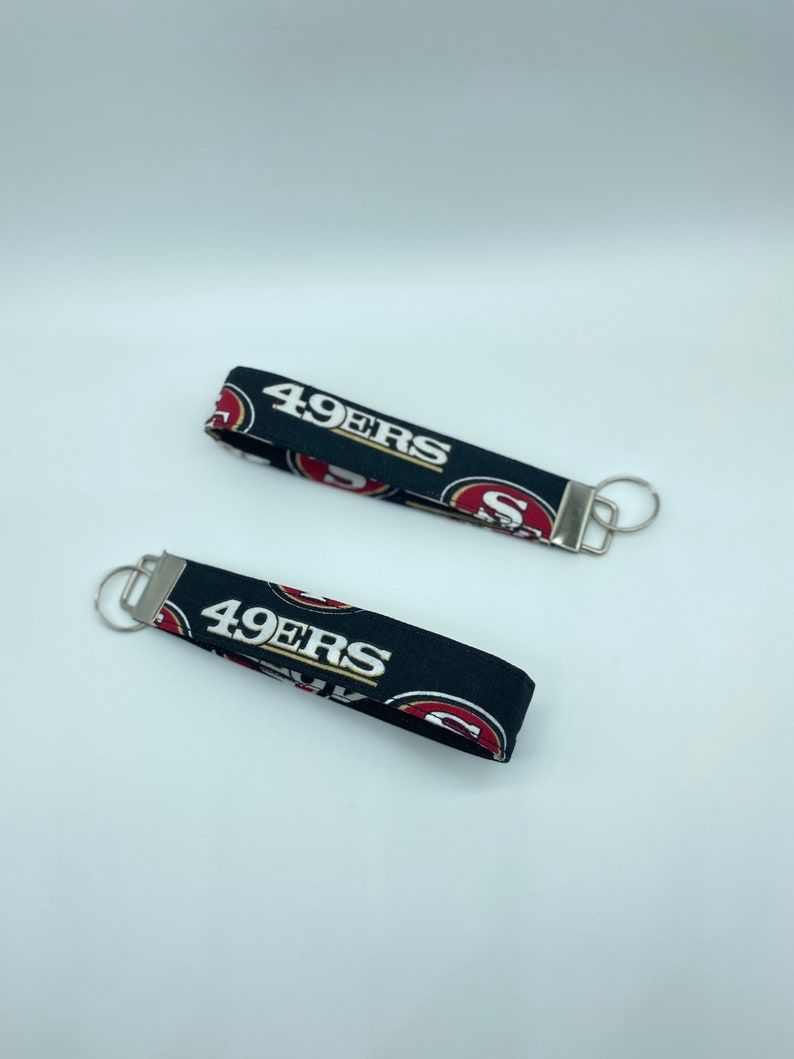 Puede incluir: Dos llaveros negros y rojos con el texto "49ers" en blanco. Los llaveros tienen un llavero plateado.
