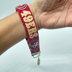 Puede incluir: Pulsera de tela roja y dorada con el texto "49ers" y un logotipo rojo y dorado. La pulsera tiene un llavero plateado.
