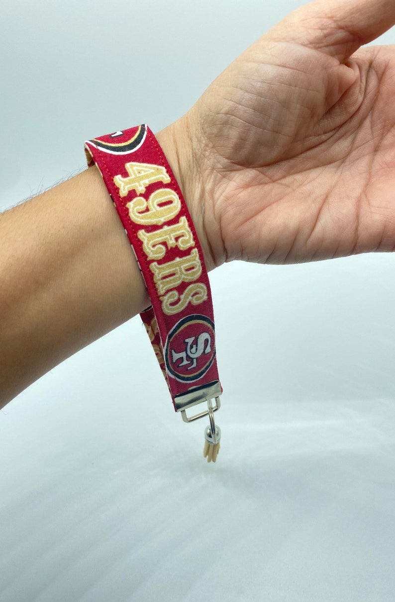 Puede incluir: Pulsera de tela roja y dorada con el texto "49ers" y un logotipo rojo y dorado. La pulsera tiene un llavero plateado.