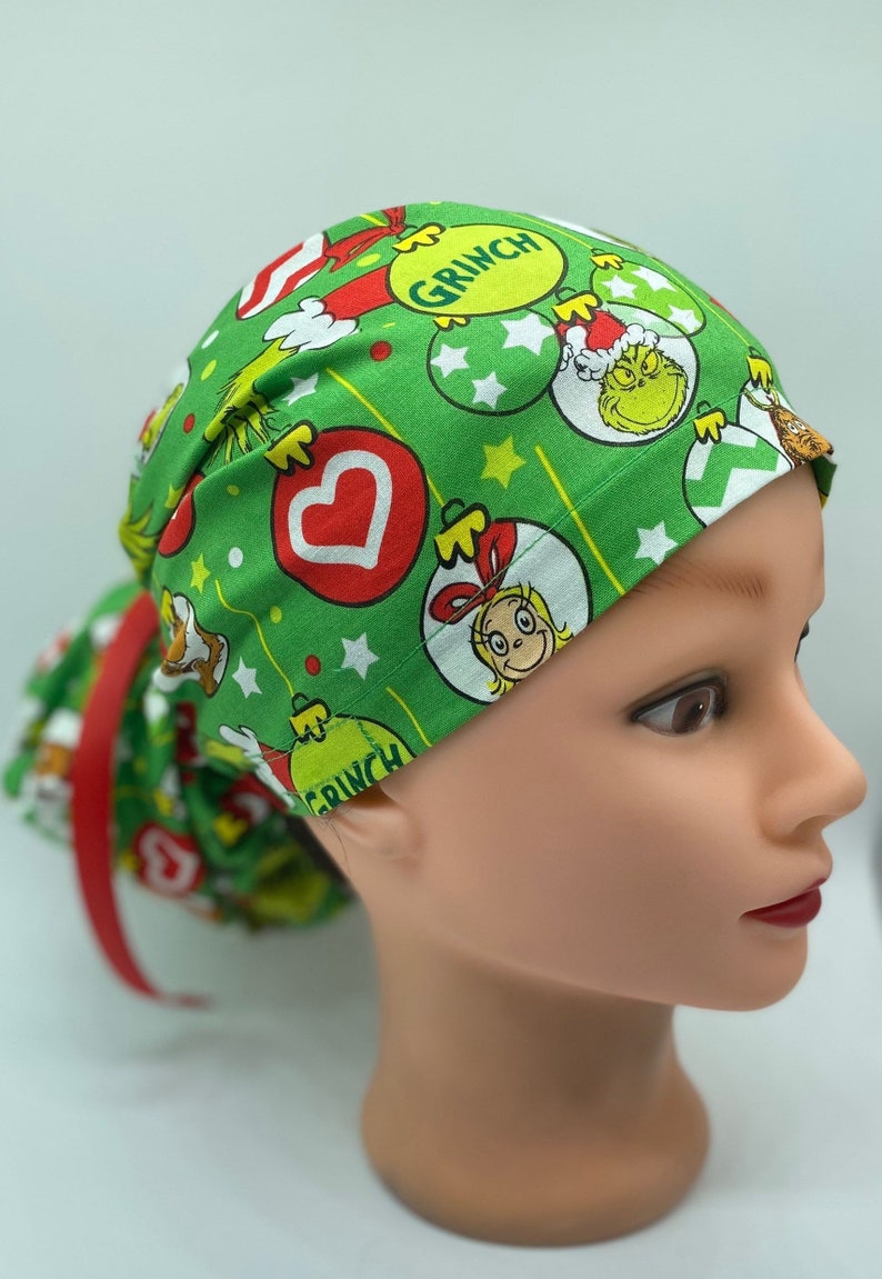 Może przedstawiać: Zielony chirurgiczny czepek z motywem świątecznym z Grinchem, Cindy Lou Who i ozdobami. Czepek ma czerwony krawat.