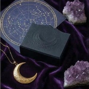 Puede incluir: Un colgante de media luna dorada con detalles intrincados descansa sobre una superficie de terciopelo morado oscuro. Una caja azul oscuro y una carta celeste están cerca, junto con dos racimos de amatistas. La escena evoca un tema místico y celestial.