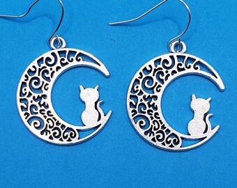 May include: A pair of silver earrings featuring a crescent moon design with a cat silhouette sitting on the moon. The moon is decorated with intricate swirls and patterns.