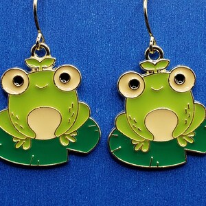 May include: A pair of gold-tone earrings featuring a green frog sitting on a green lily pad. The frog has large, white eyes with black pupils and a small, green leaf on its head.