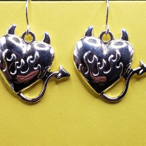May include: A pair of silver dangle earrings featuring a heart shape with flames and devil horns and tail.