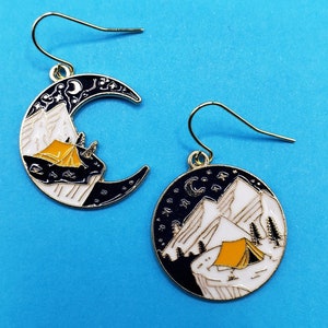 May include: A pair of gold earrings with a crescent moon design. One earring features a black and white mountain scene with a yellow tent, while the other earring features a black and white mountain scene with a yellow tent and stars.