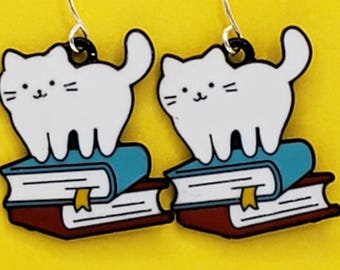 May include: A pair of cat-themed earrings. Each earring features a white cat with black accents standing on a stack of books. The books have blue and maroon covers with a yellow bookmark. The earrings hang from silver-colored hooks.