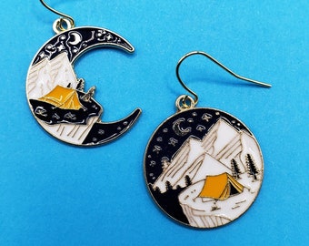 May include: A pair of gold earrings with a crescent moon design. One earring features a black and white mountain scene with a yellow tent, while the other earring features a black and white mountain scene with a yellow tent and stars.