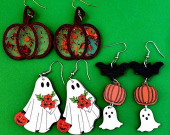 May include: A collection of Halloween-themed earrings. The earrings include pumpkin-shaped designs with red and orange accents, white ghost earrings with red flower bouquets, and bat and pumpkin designs. The earrings are set against a green background.