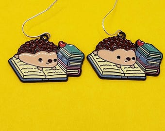 May include: A pair of whimsical earrings featuring a cartoon hedgehog reading a book. The hedgehog has brown quills and a tan body, and is lying on an open book. A stack of books with a red heart on top is next to the hedgehog. The earrings have silver hooks.