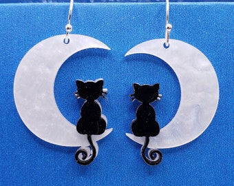 May include: A pair of silver earrings with a crescent moon design. Each moon has a black silhouette of a cat sitting on it.