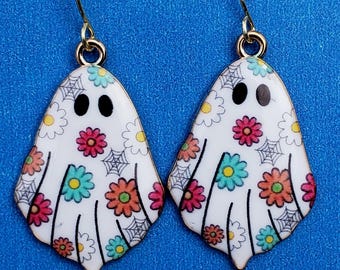 May include: A pair of ghost-shaped earrings with gold-colored hooks. The white ghosts are decorated with colorful flowers, black spiderwebs, and black oval eyes. The earrings are set against a blue background.