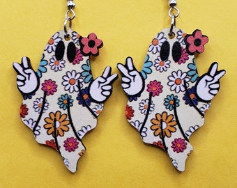 May include: A pair of ghost-shaped earrings featuring a cream-colored base with a multi-colored floral pattern, black oval eyes, and white hands making peace signs. Each ghost has a small pink flower accent near the top and silver hooks.