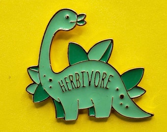 May include: Green enamel pin of a cartoon dinosaur with the word "Herbivore" written on its body. The dinosaur has a long neck and a spiky back.