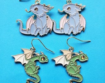 May include: Four pairs of dragon earrings. Two pairs are light blue with pink wings, and two pairs are green with white wings. All have gold-colored hooks and are set against a bright blue background. The earrings feature a cartoon-style design.